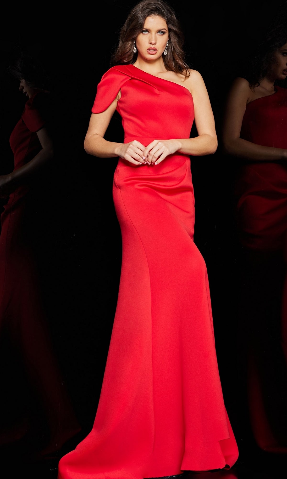 A woman stands against a black background, hands clasped, wearing the Jovani 06753 Long Formal One-Shoulder Dress with her loose hair styled in waves.